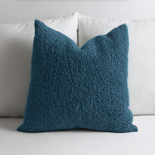 A blue, textured throw pillow sits on a white bed, contrasting with the white pillows behind it.